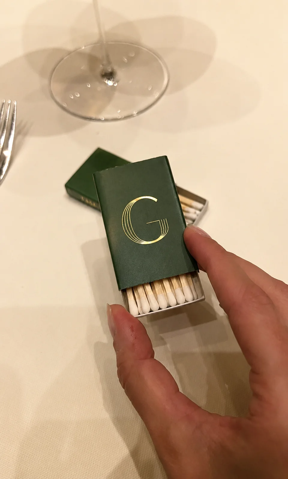 A dark green matchbox with a gold monogram, held above a restaurant table