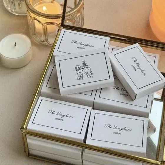 Cajas de cerillas de boda personalizadas con nombres dibujados a mano, apiladas en una caja de cristal